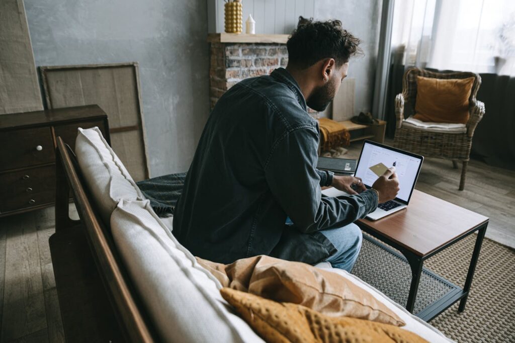 A man wearing a denim jacket makes an online purchase using his laptop while sitting indoors on a couch.